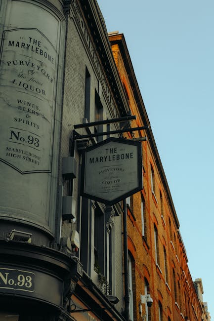 An exterior view of a historic building on Marylebone High Street, featuring two black-framed, rectangular signs hanging from the facade that read 'The Marylebone' and 'Pure Victorian Liquor.' The building's facade is made of red and beige brick, with black window frames. The signs are mounted at different heights, with one slightly tilted, against a clear blue sky. The scene reflects the character of the Marylebone district, known for its traditional shops and heritage architecture, relevant for interior cleaning and surface maintenance discussions related to residential and commercial properties in the area.