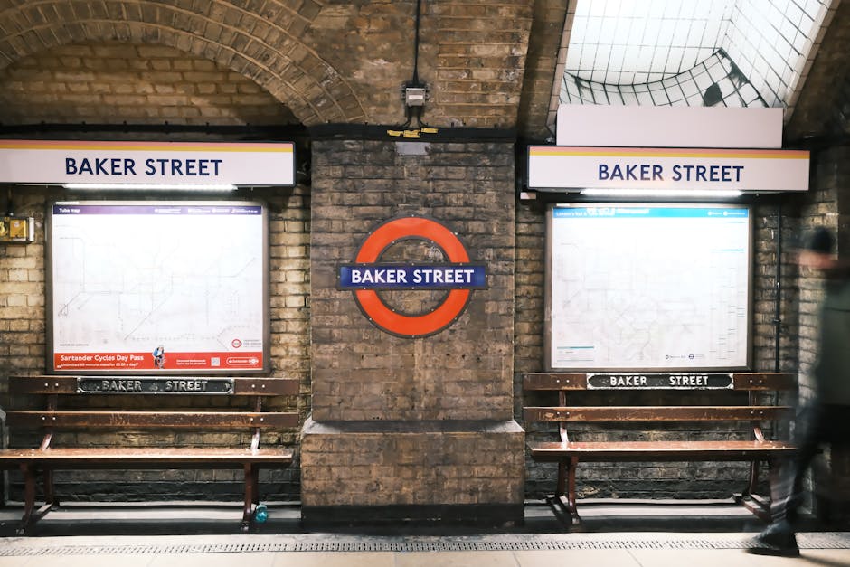Interior view of Baker Street underground station featuring two wooden benches beneath illuminated route maps protected by glass, with a prominent red circular Baker Street station sign mounted on a brick wall between the maps. The station’s brick arches and tiled ceiling are visible, with soft lighting illuminating the scene. A blurred person walks past on the platform, highlighting the busy yet clean environment. Marylebone Cleaner offers comprehensive domestic cleaning services, ensuring surfaces such as these are meticulously cleaned and sanitized to maintain hygiene and appearance in residential and commercial spaces.