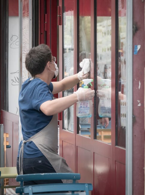 The exterior of a retail shop on Marylebone High Street featuring a large glass display window and a dark wooden frame. Several cleaning tools, including brooms with natural straw bristles, dusters, a